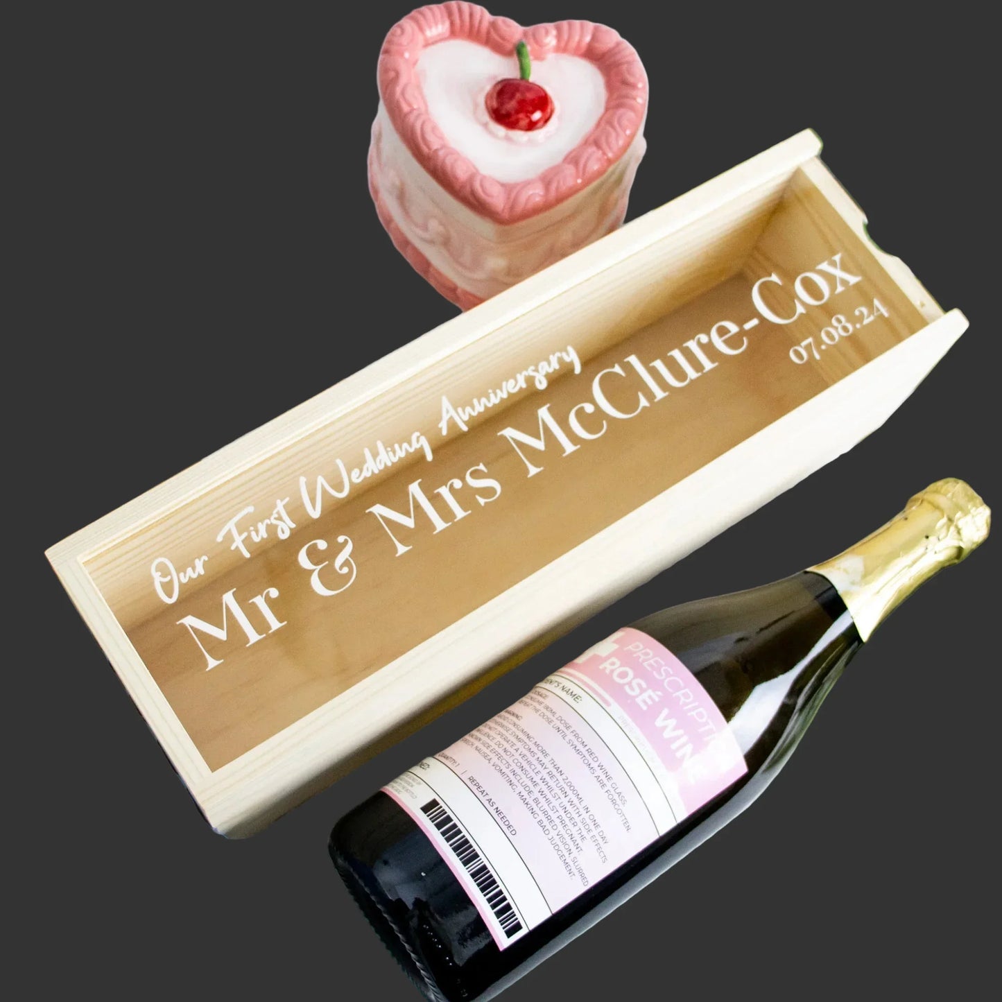 Printed wooden wine and champagne box with engraved wedding anniversary text, a wine bottle, and a pink heart-shaped cake on a dark background.