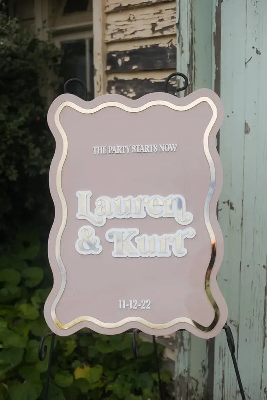 Decorative wedding welcome sign for Lauren and Kurt with event date, outdoors by rustic building
