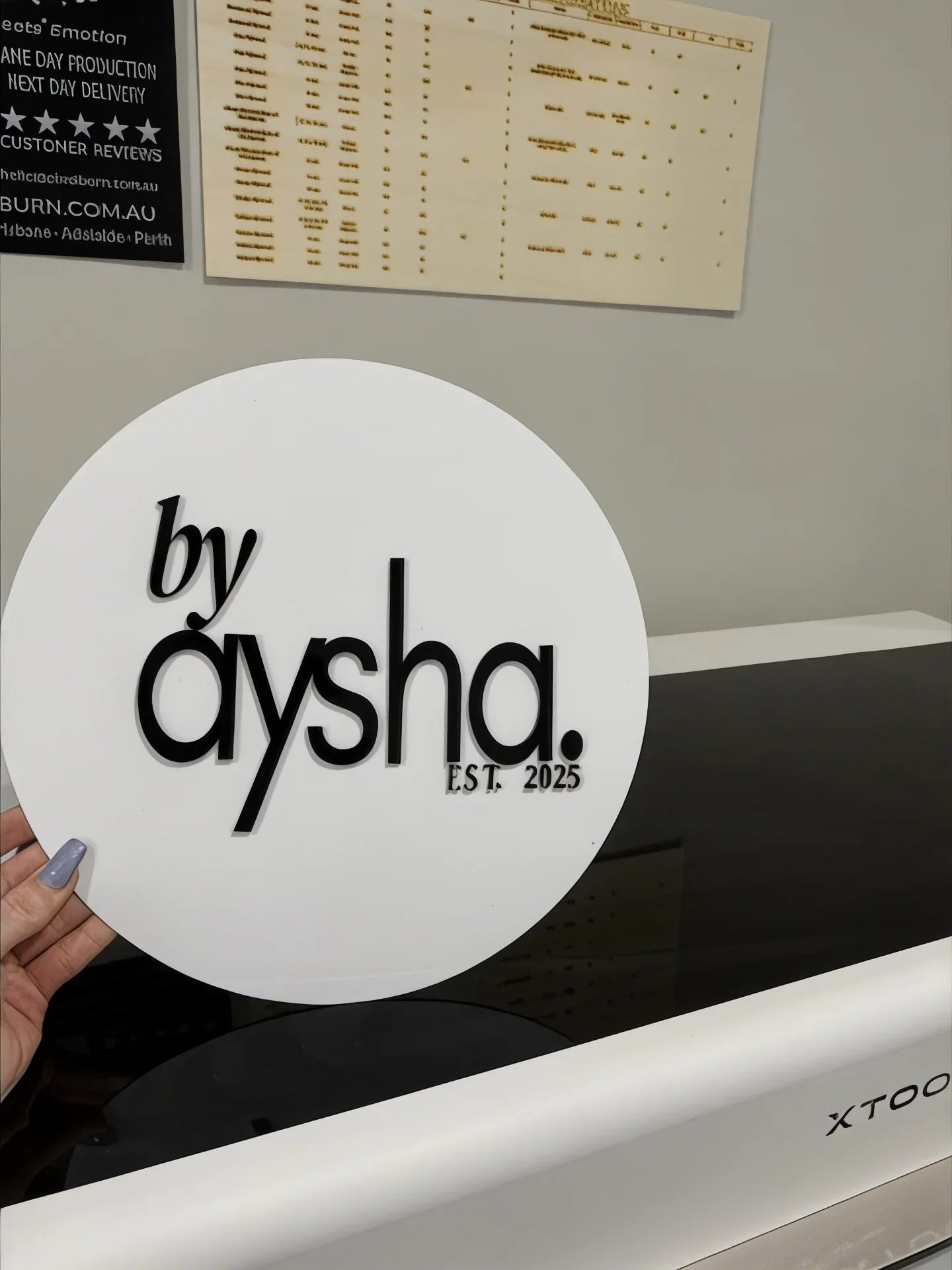 Hand holding custom round acrylic sign by aysha EST. 2025 in workshop setting