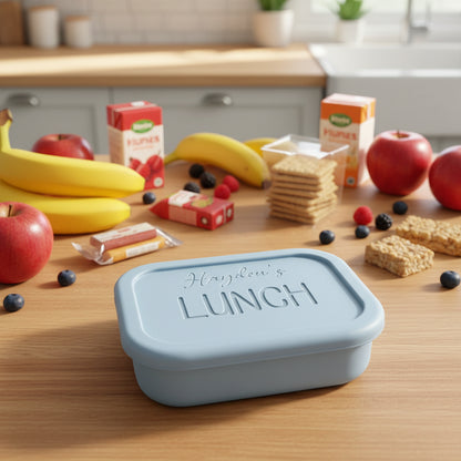 Light blue lunch box with 'Hayden's LUNCH' text on a wooden surface with fruits and drinks.